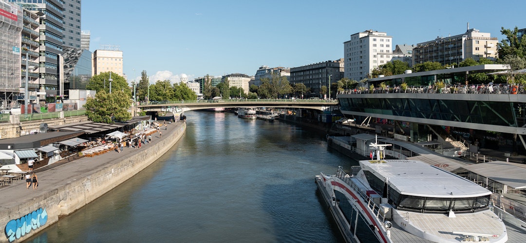 A narrow waterway winds its way through an urban environment, with a large boat docked on the side. It's sunny and bright, with trees in the background. 