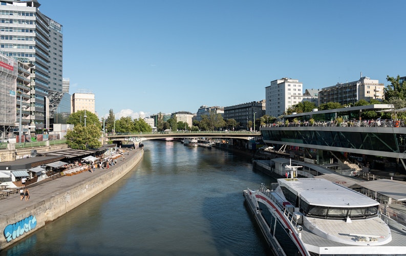 A narrow waterway winds its way through an urban environment, with a large boat docked on the side. It's sunny and bright, with trees in the background. 