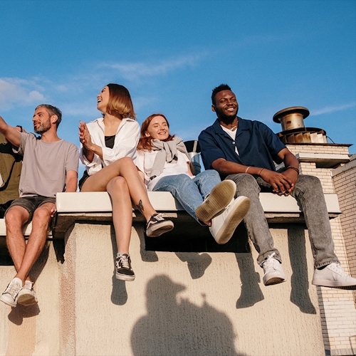 Four young people sit on a raised stone surface, smiling and pointing. They're squinting into the day's fading light. 
