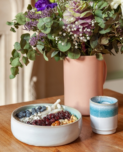 A shallow, white ceramic bowl sits on a wooden table in front of a vase with flowers. The bowl holds a variety of small fruits and nuts. 