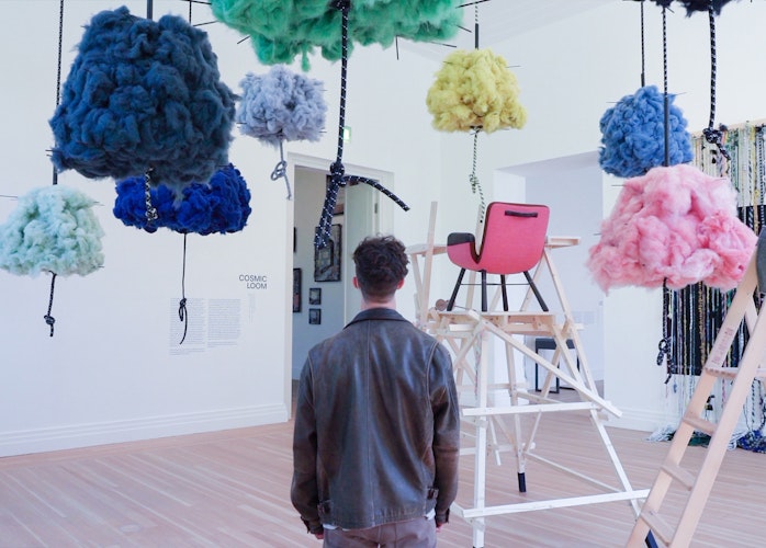 A young man stands in the middle of an art exhibition featuring colourful clouds hanging from the ceiling