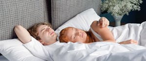 A photo of a young white couple lying awake in a white bed
