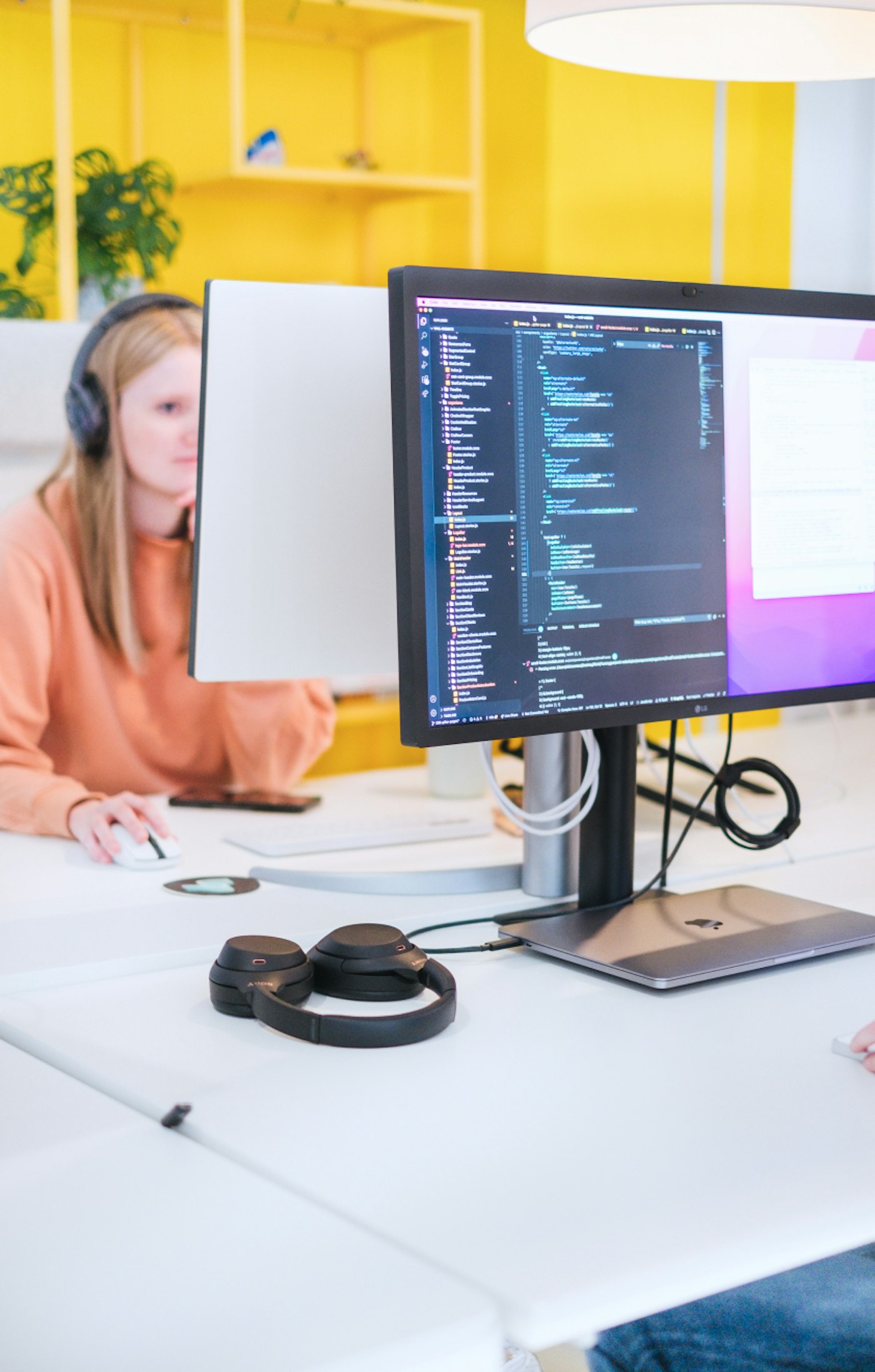 Code editor shows on a monitor while development intern Margot shows out of focus in front of a yellow office wall.