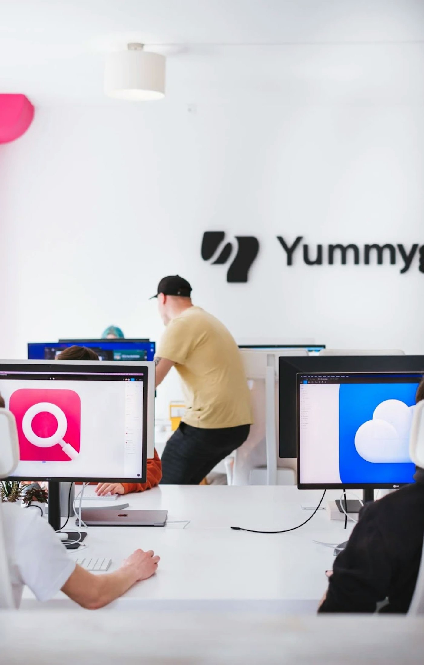 Two designers as seen from the rear who are looking at their monitors that show zoomed in colorful icons with person further in image hovering over collegeagues chair. Dark antracite Yummygum logo on an office wall  is centered in the very rear.