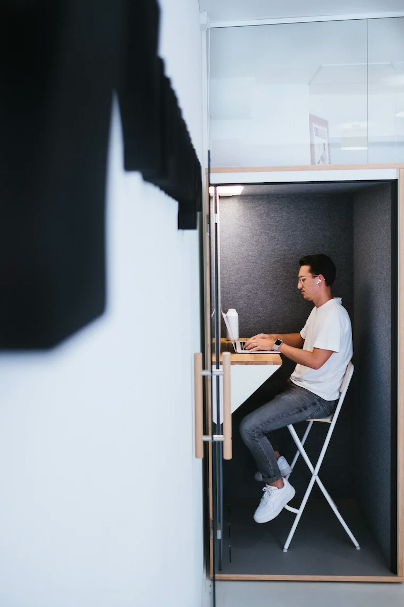 Yummygum co-founder Vince sitting on a bar stool working on his laptop. The bar stool stands in an acoustic phone booth. Cropped the foreground, partly out of focus is the Yummygum embossed logo on a white office wall.