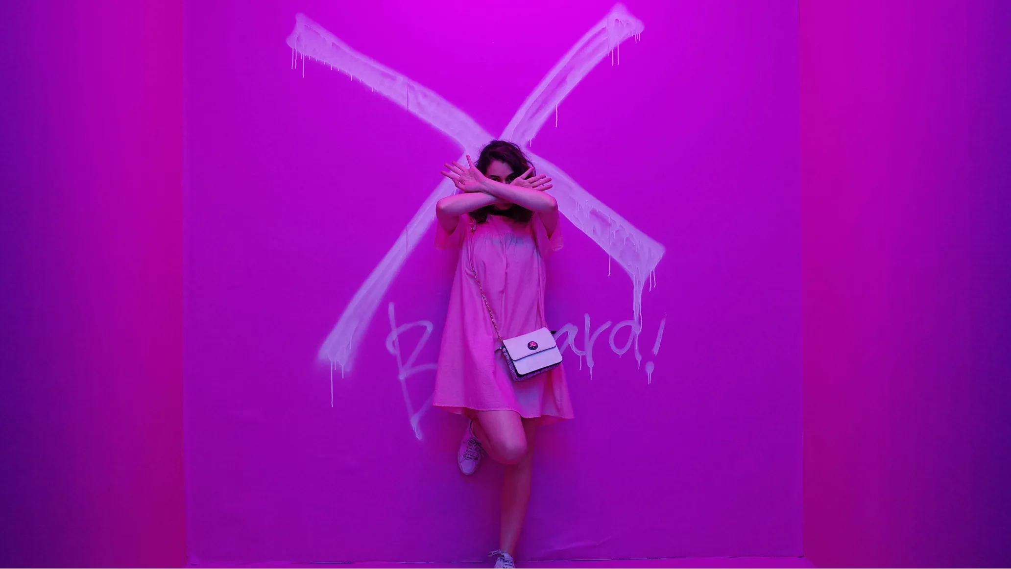 female with arms crossed in pink dress in front of a pink purplish wall that shows a large cross painted dripping graffiti paint suggesting