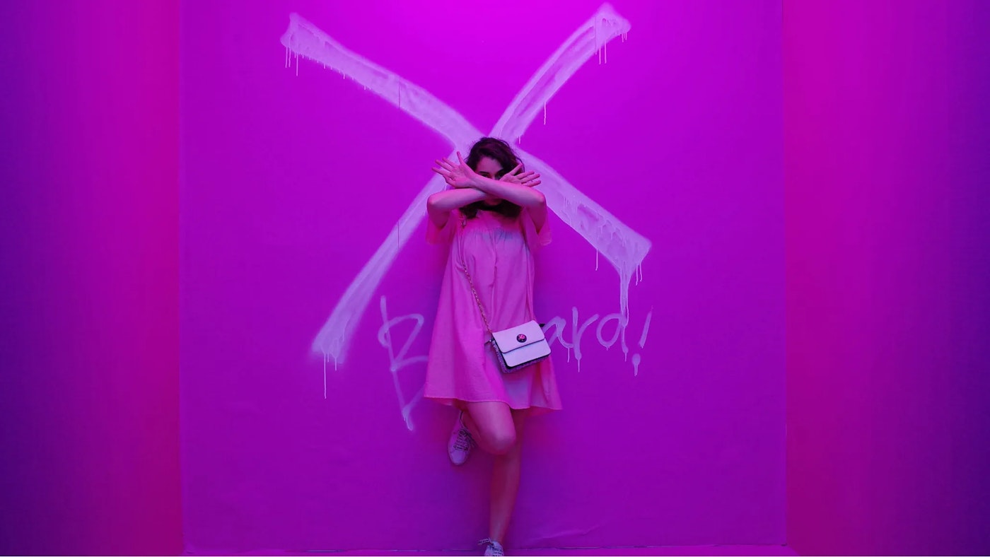 female with arms crossed in pink dress in front of a pink purplish wall that shows a large cross painted dripping graffiti paint suggesting