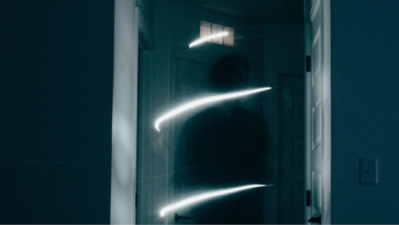 A mysterious looking shade of what looks like a human in front of a low lit hallway. The hallway is ever so slightly lit by three dashes of light that are obscuring the person shade.