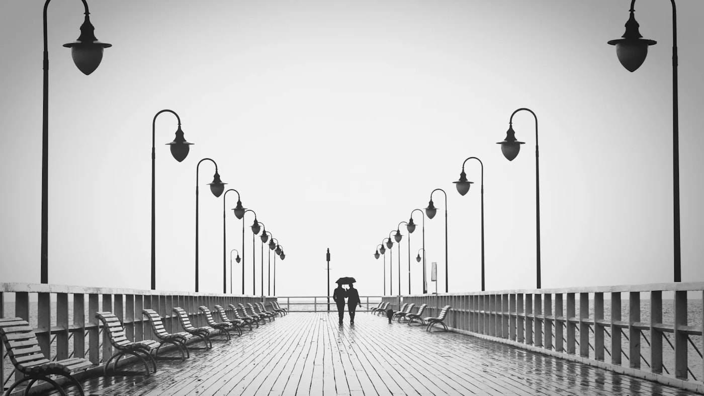 Black and white symetric scene of an empty and rainy board walk with benches and lanterns on each side , with in the center a couple walking their dog and sharing an umbrella.