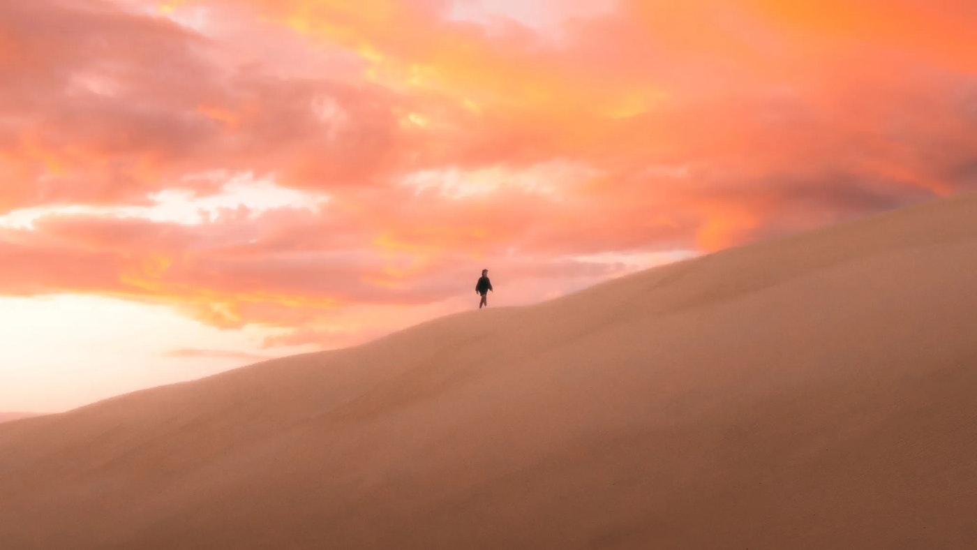 Silhouette of a person walking on a desert mountain ridge with an cloudy orange pink sky as a backdrop.