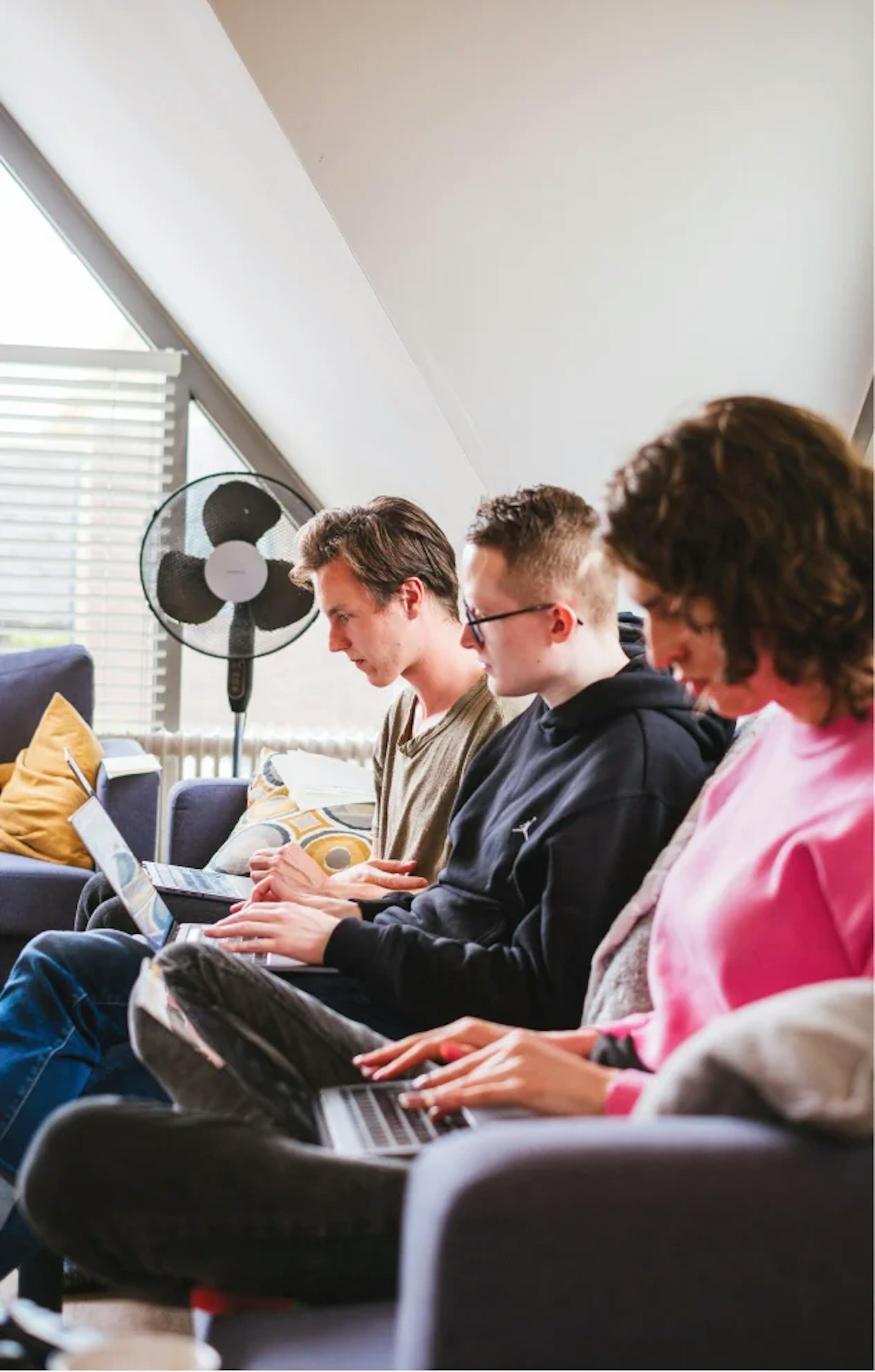 Three people sitting side by side on a couch all working on their laptops, as seen from the side