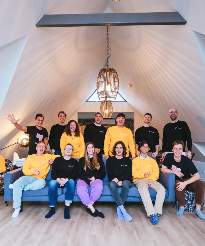 Team photo of people who work at Yummygum, of which the half is sitting on a couch and the rest is standing behind the couch, all looking at the camera. Some are wearing black Yummygum swaters, others are wearing yellow Yummygum sweaters