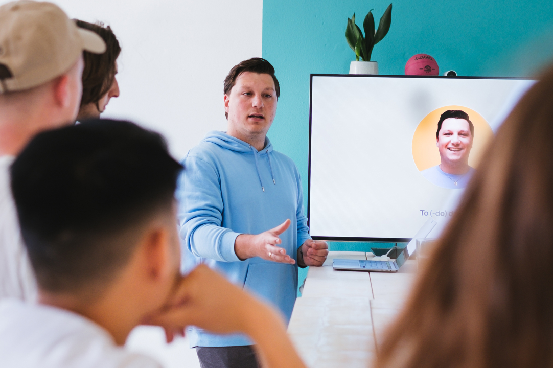 Person giving presentation guided by a slide deck displayed on a large monitor. Person is seen over the shoulders of people listening to the presentation