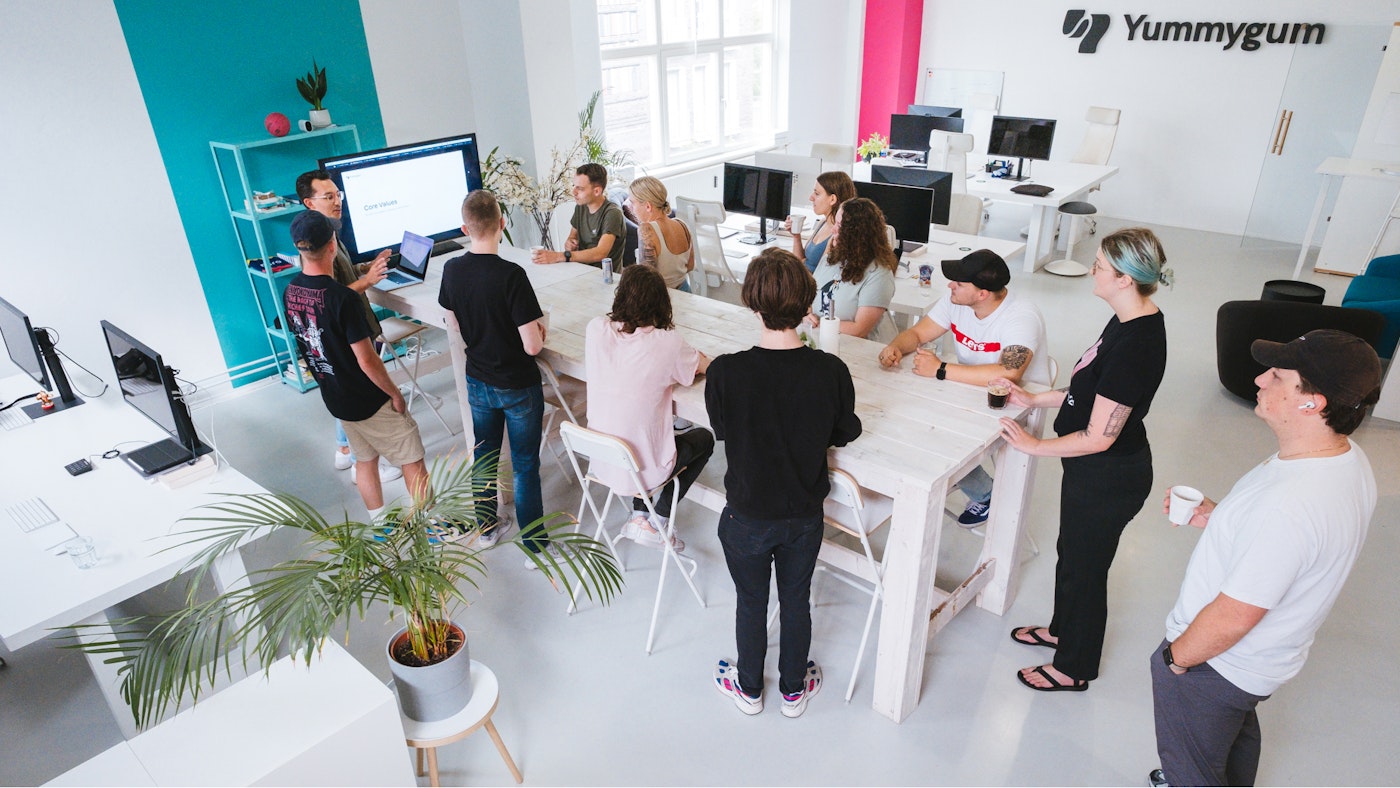 A group of people, the Yummygum team, standing in a minimalist white office space. Everyone is standing around a large pub table. One person is giving a presentation on a large monitor. The rest of the group is listening and watching the presentation.