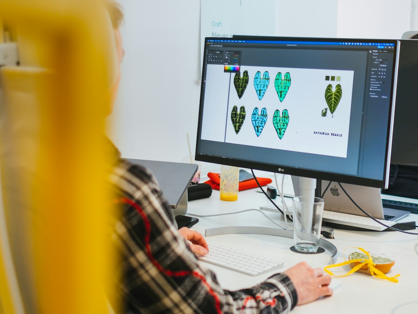 Person working on a computer display as seen from the back. The display shows a dark user interface and a white canvas which contains leaf shaped illustrations.