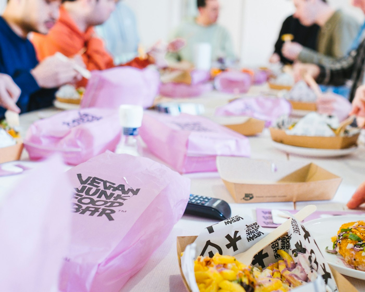 Pink paper packaging from Vegan Junk Food Bar on a table that also holds plates that have cardboard trays containing vegan fast food snacks.