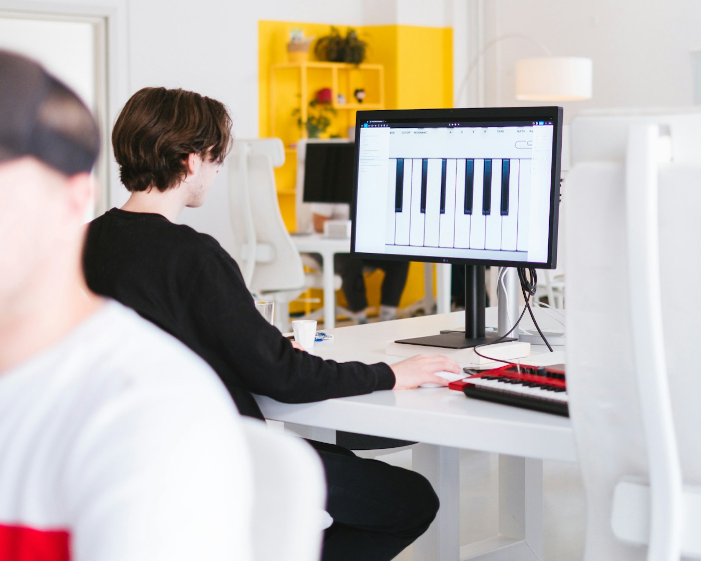 Person sitting behind a display that shows a close up of synthesizer keys as recreated using user face web application Figma. Besides the person on the desk a synthesizer shows.