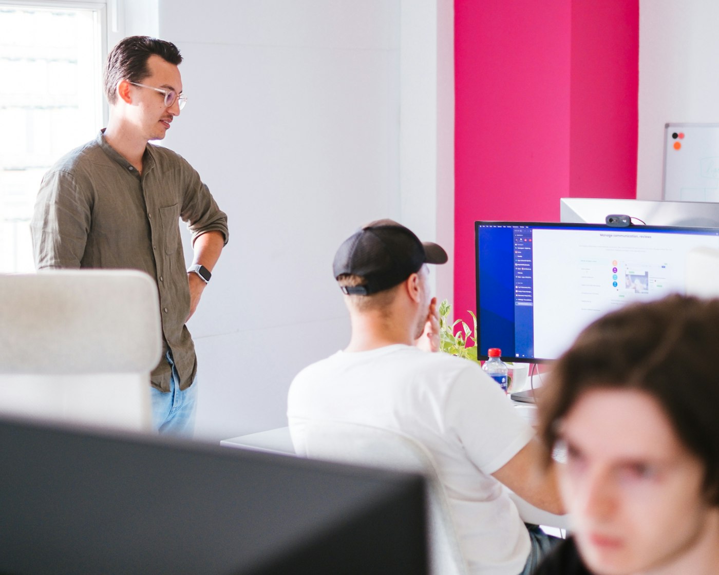 Person standing besides other person's desk as they seem to actively talk about what is showing on a monitor. In the rear there is a narrow bright pink wall, contrasting against the other, white walls.