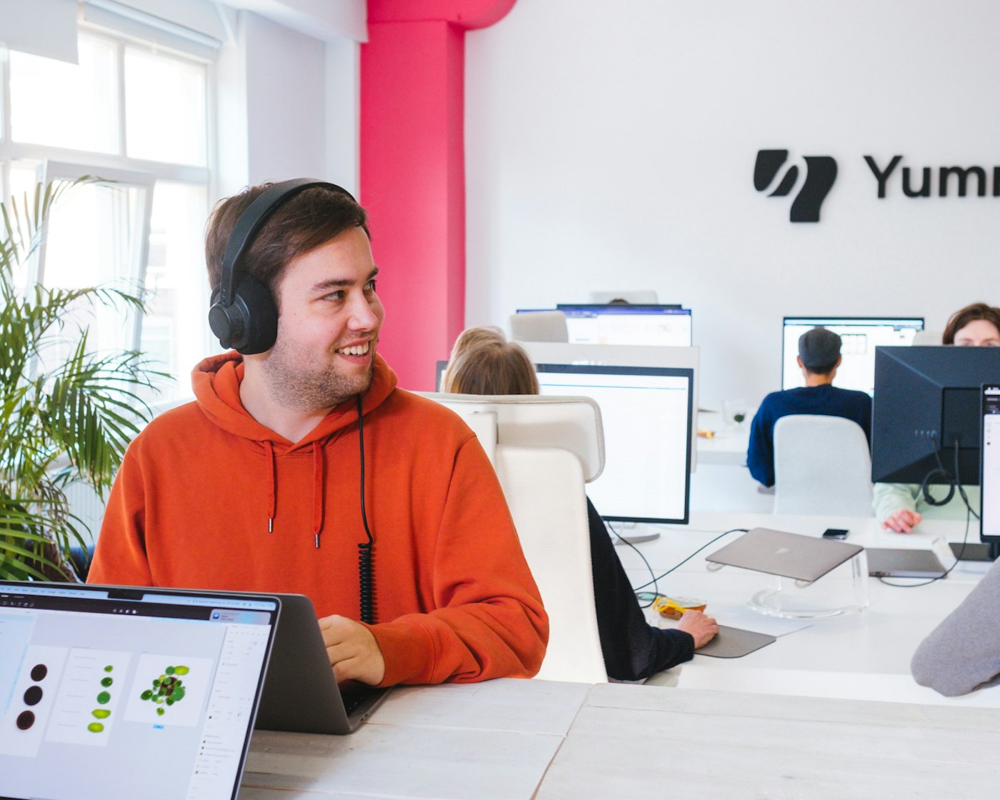 Person in orange red hoodie with head phones on their head looking side ways as they are standing behind a laptop. In the back are people sitting behind their desks..