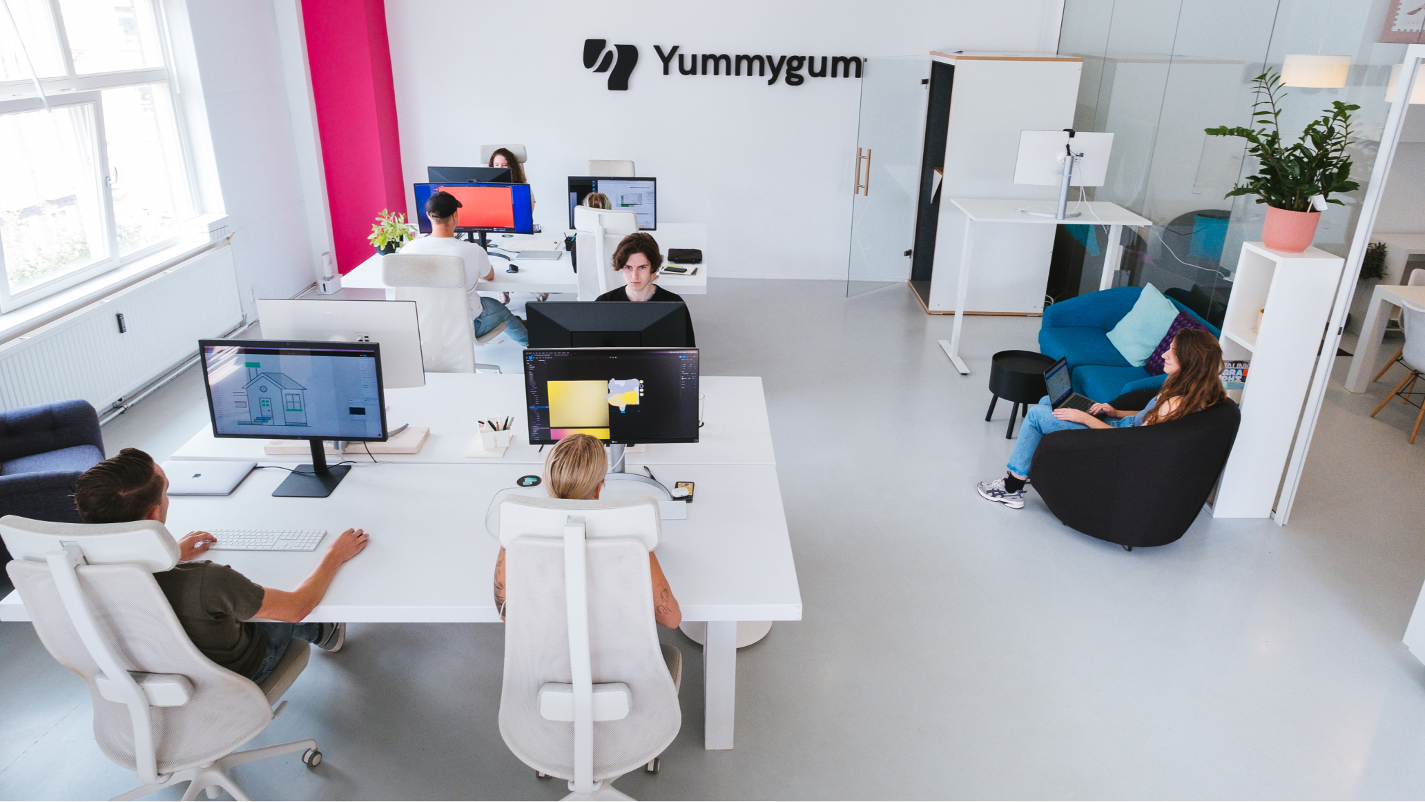 Wide angle top down view of the clean and minimalist Yummygum office that shows a black embossed logo that says Yummygum. The office shows two groups of two white desks with people working behind computers. Another person is actively working on their laptop as they sit in a dark anthracite chair.