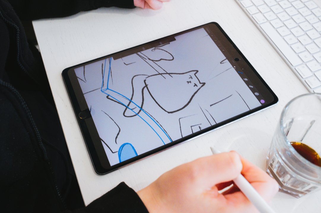 Ipad on a white desk with two hands and a near empty coffee cup beside it. The ipad shows an illustration of a cat. The right hand is holding a white ipad stylus.