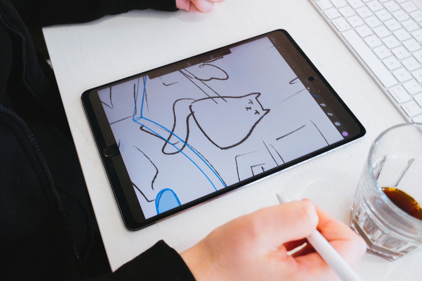 Ipad on a white desk with two hands and a near empty coffee cup beside it. The ipad shows an illustration of a cat. The right hand is holding a white ipad stylus.