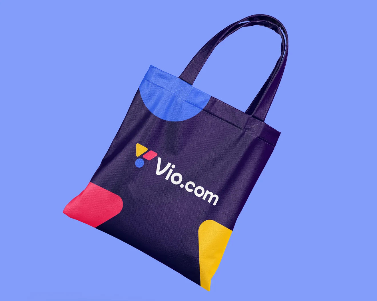Tote bag on a light blue background. The tote bag has a dark blue color and shows  the Vio.com logo with a white logotype combined with a white logo mark consisting of three colored shapes. On three of the four tote bag's corners are large partially cut off colored shapes
