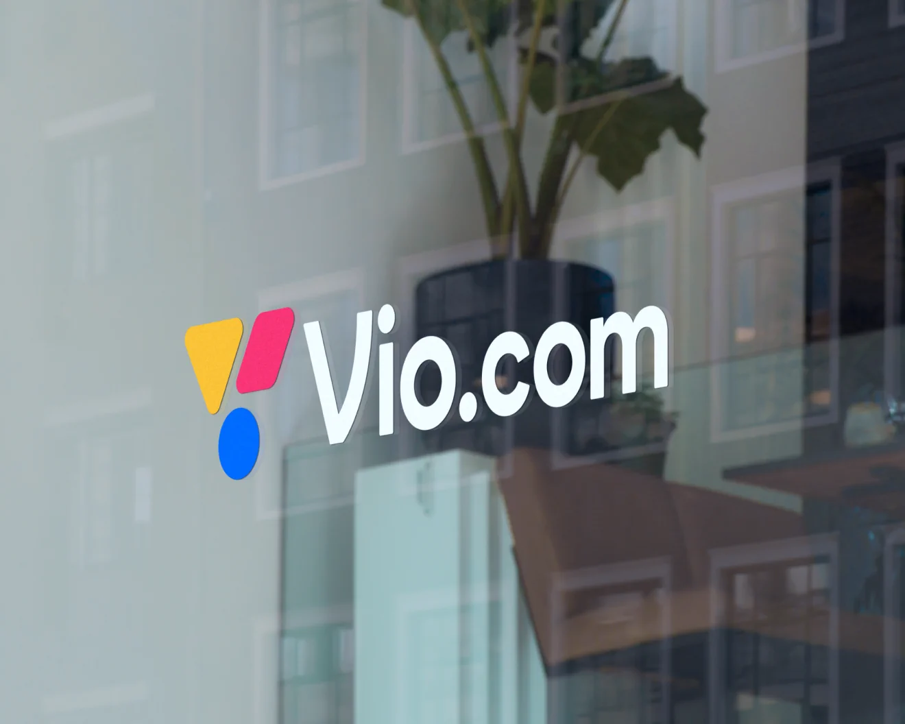 A photo of a glass window that shows the Vio.com logo in white text with yellow, blue and magenta-red colored shapes next to it. Behind the window is a plant and a couch, seemingly an office space.