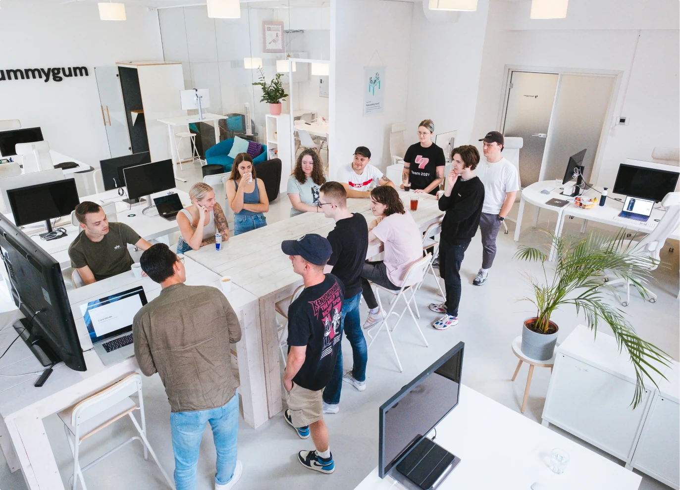 Group of people standing and sitting around a bar table in a bright office. Most of the people are looking in the direction of a person standing besides a large television, seemingly giving a presentation