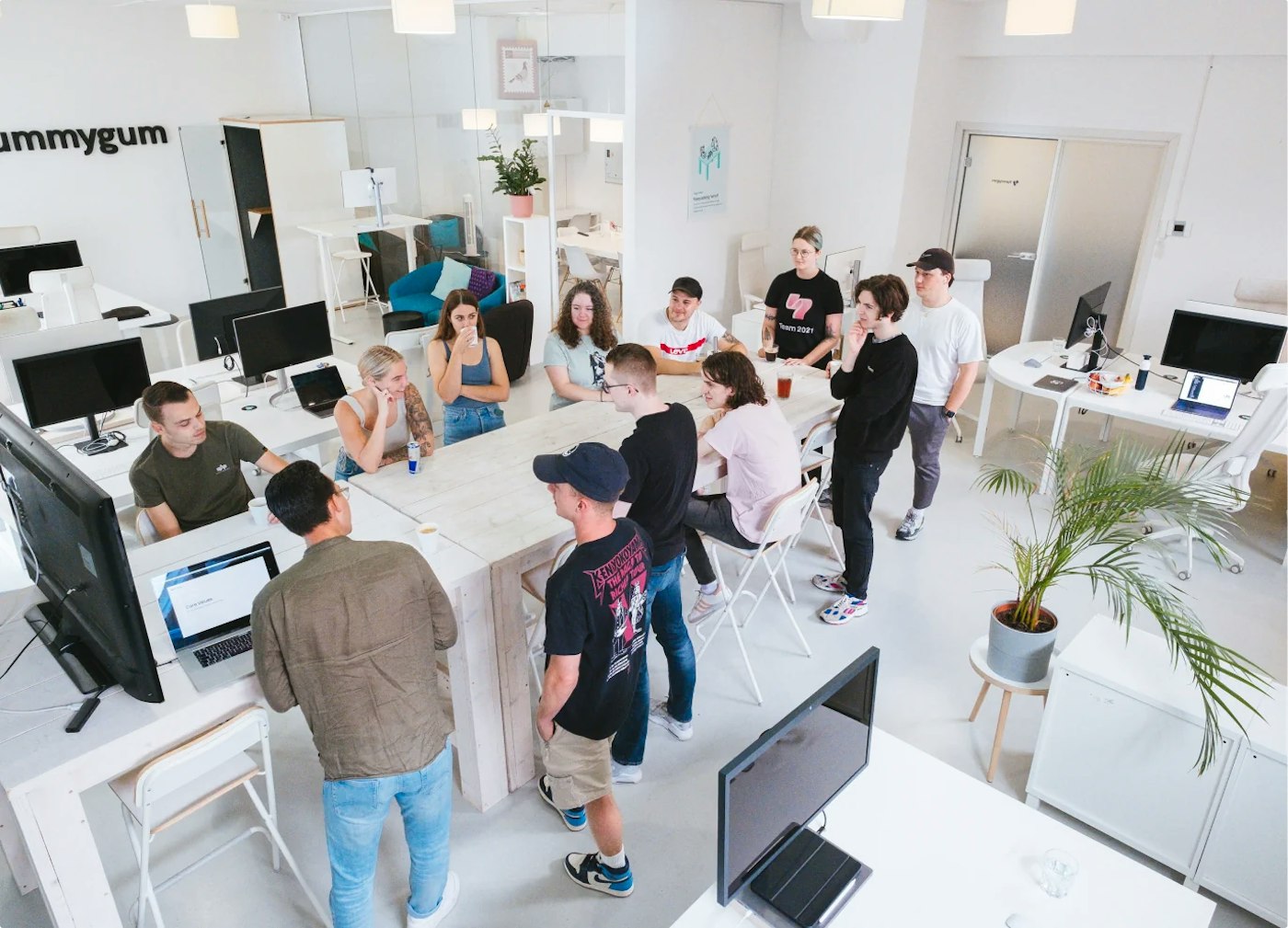 Group of people standing and sitting around a bar table in a bright office. Most of the people are looking in the direction of a person standing besides a large television, seemingly giving a presentation