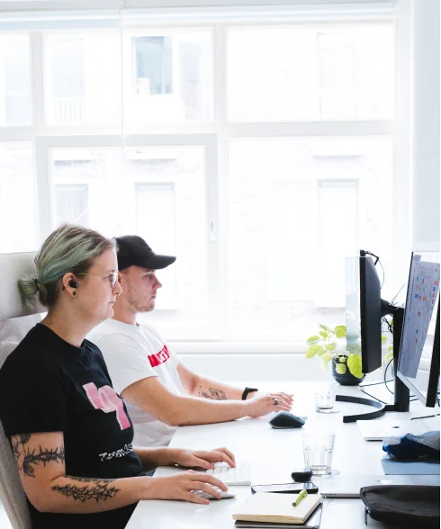 Two people as seen from the side, sitting behind their joined desk, working side by side. In the back are bright window panes.