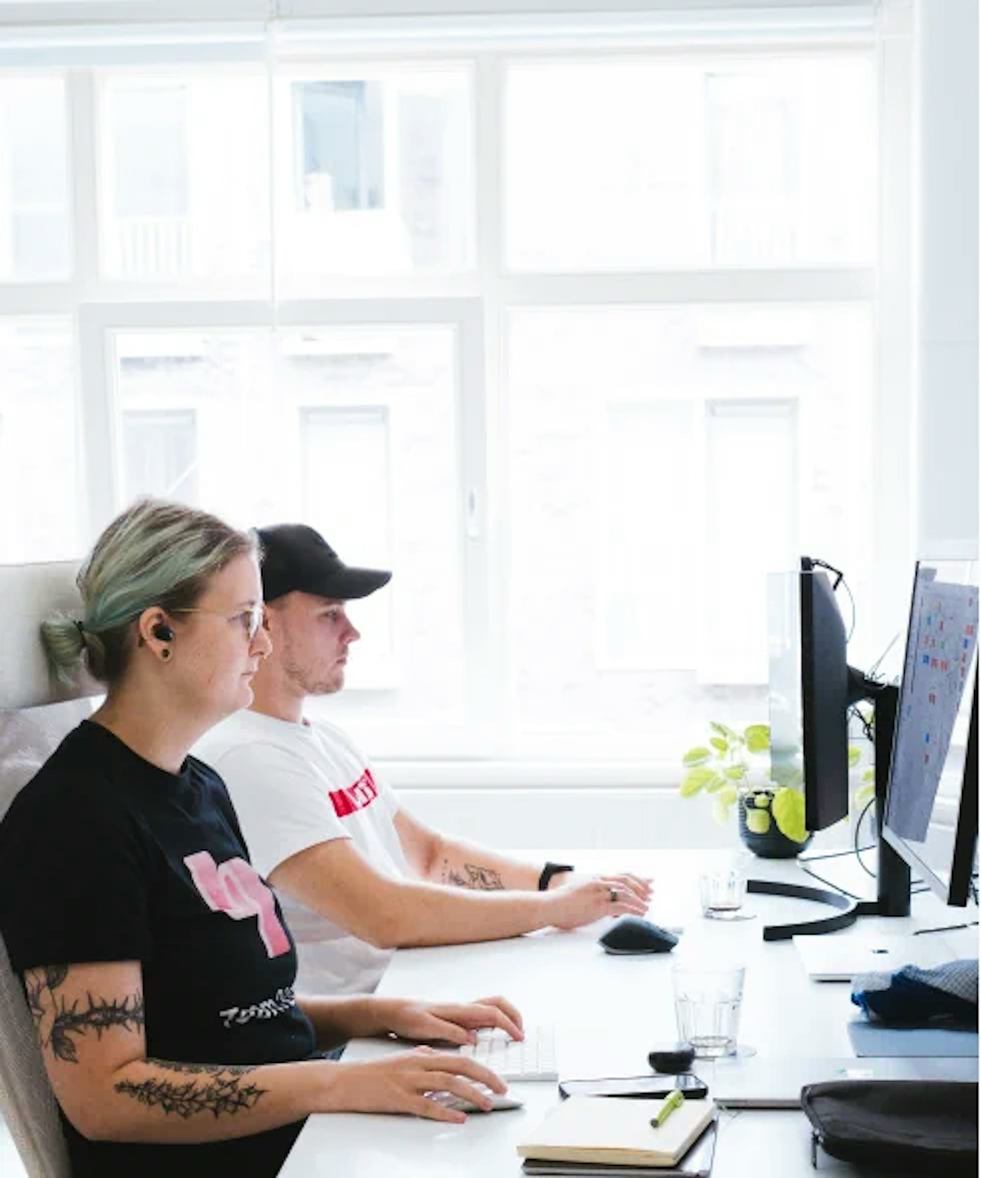 Two people as seen from the side, sitting behind their joined desk, working side by side. In the back are bright window panes.