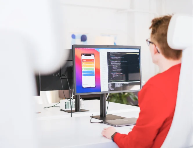Person with red sweater, as seen from the rear sitting behind a desk. Their monitor shows a code editor and a mobile app interface.