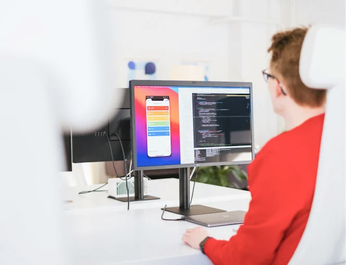 Person with red sweater, as seen from the rear sitting behind a desk. Their monitor shows a code editor and a mobile app interface.