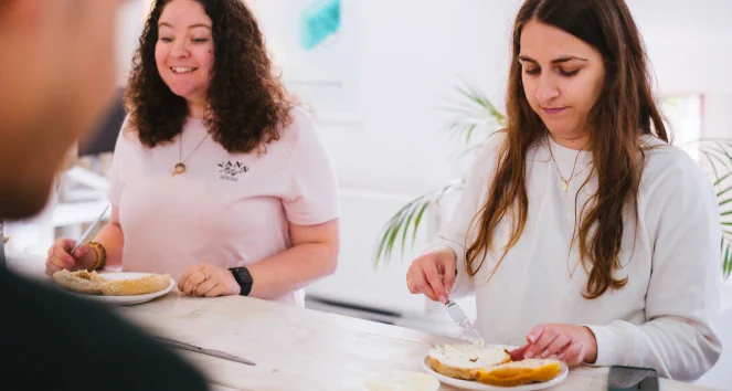 Two people, of which one is laughing, standing behind a bar table. Both are making a bagel.