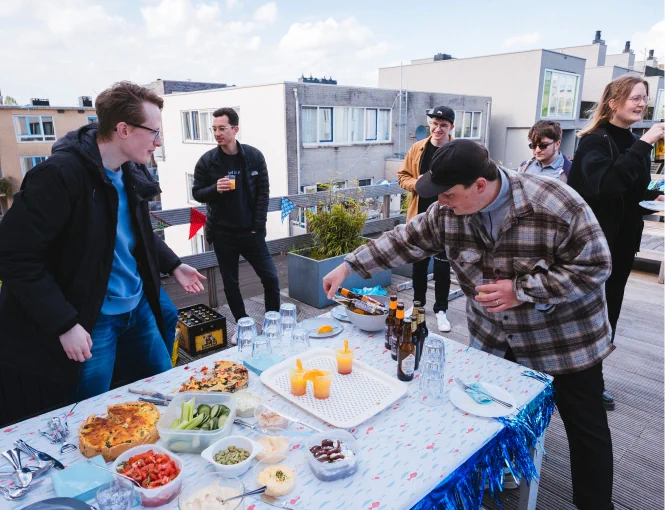Group of people at a casual company party on a rooftop. Two people hover over a table that has all kinds of foods and drinks. In the background are other buildings' rooftops.