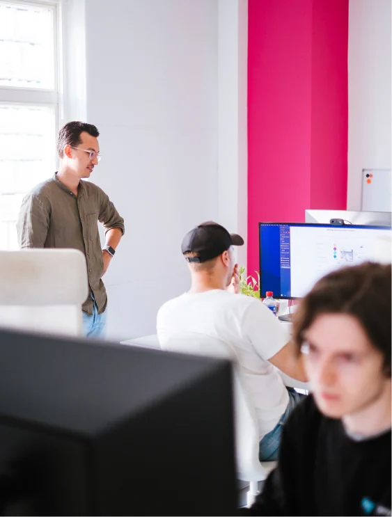 Person standing beside other person's desk. Both of them are looking at the sitting person's monitor. They are in a white office that has a bright magenta-pink painted wall.