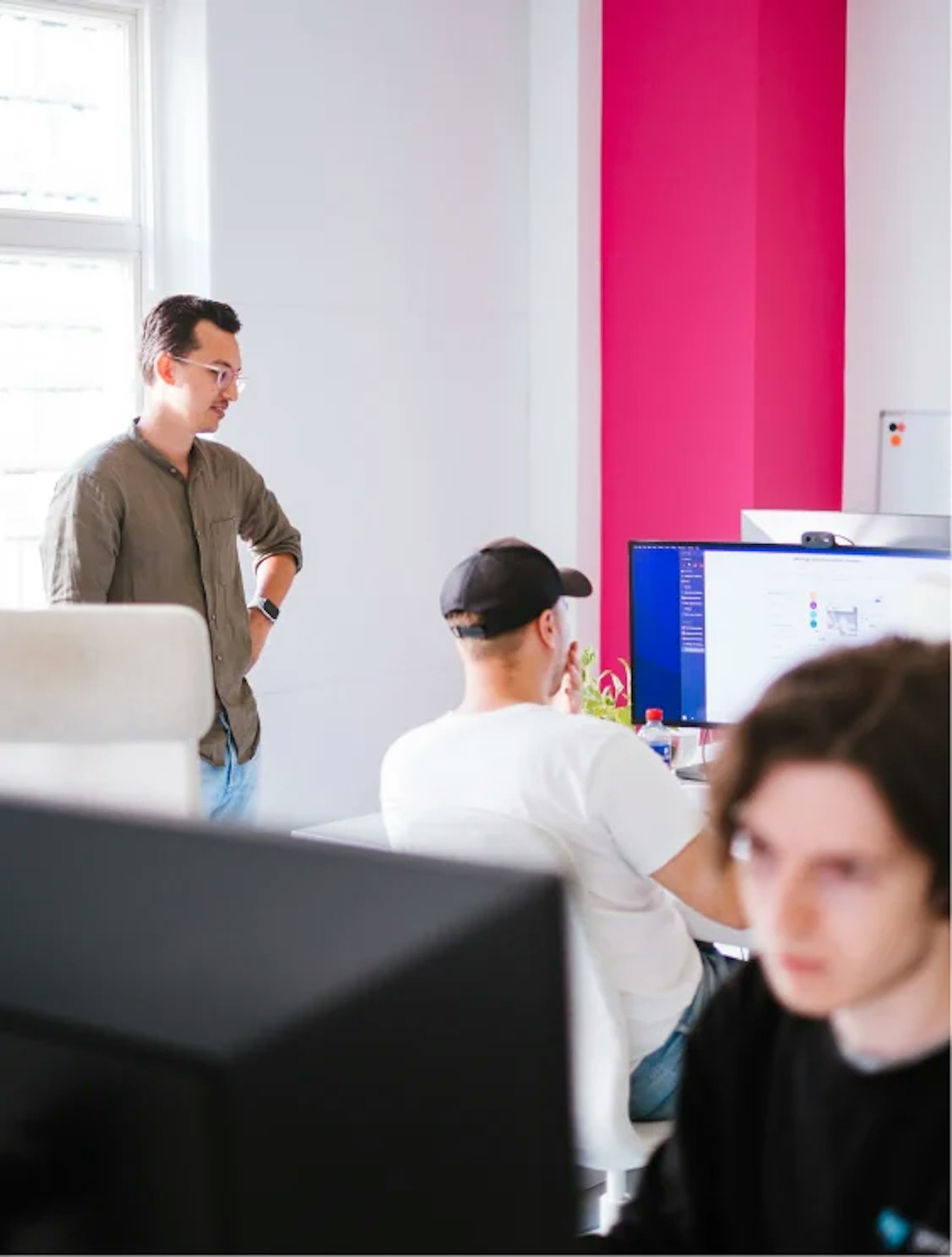 Person standing beside other person's desk. Both of them are looking at the sitting person's monitor. They are in a white office that has a bright magenta-pink painted wall.