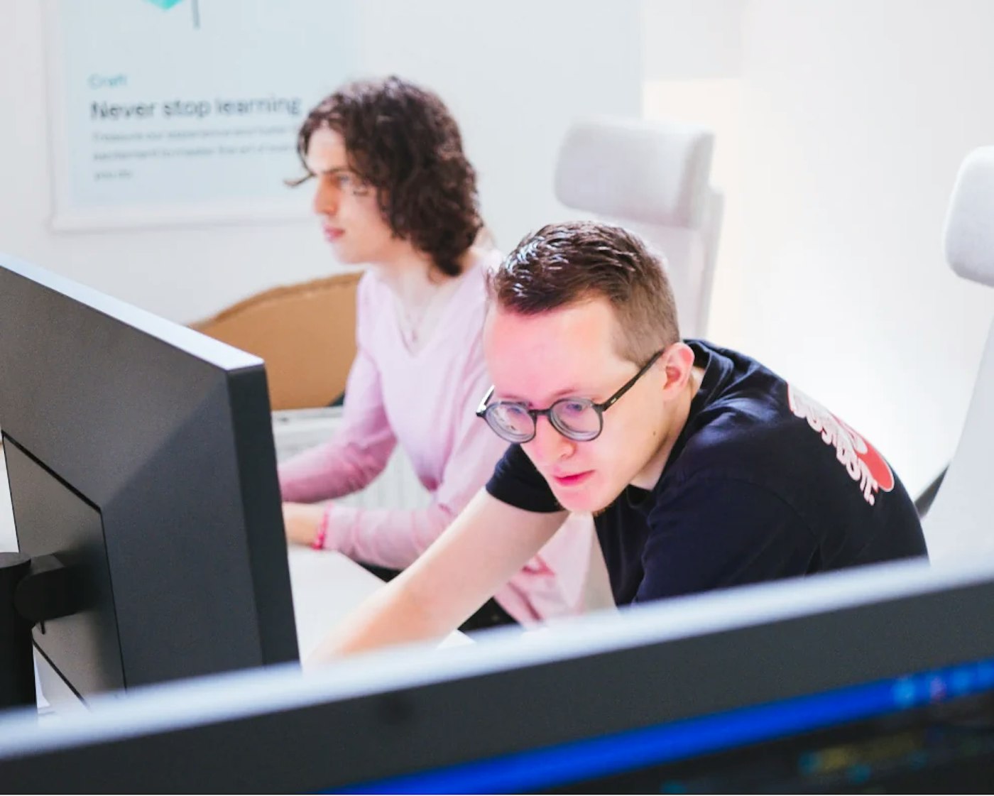 Two people sitting side by side, of which the rear person is looking at the monitor in front of them. The front most person is hunched over and looking at another monitor and seemingly strongly focused.