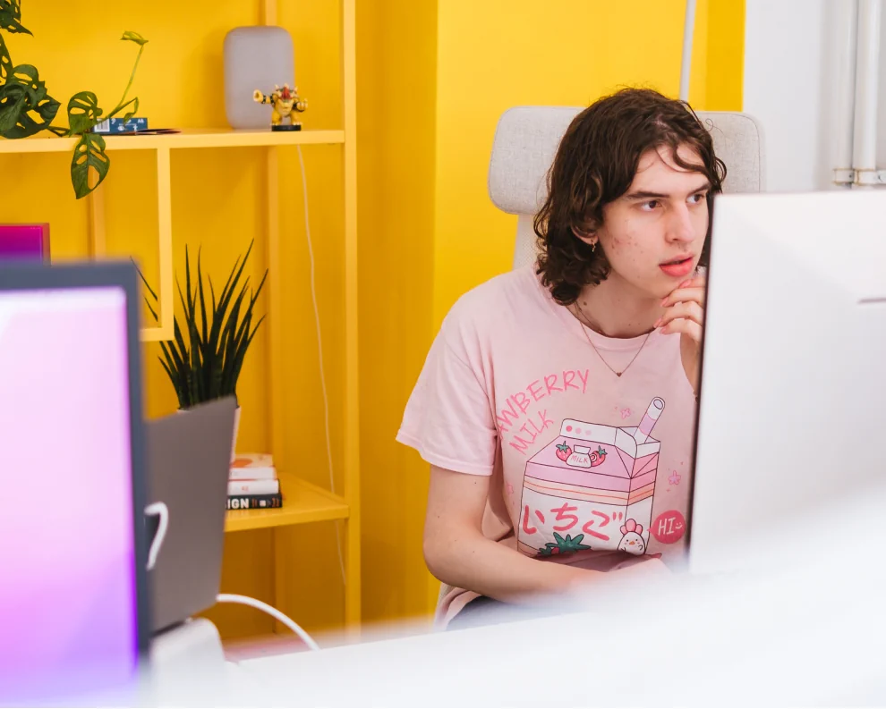 Person, as seen through a group of monitors, dressed in a pink t shirt with a cartoon on it looking at their screen while picking at their chin. Behind the person is a yellow painted wall.