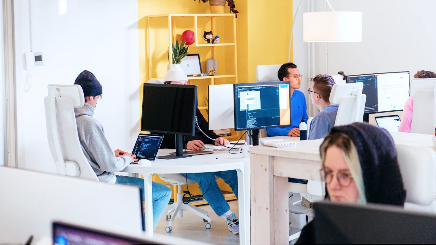 Group of people working at their desks at an office. Their monitors show code editors. In the rear there is a yellow painted wall. Closer to the person taking the picture is a person with a dark hoodie on who looks at a monitor in front of them.