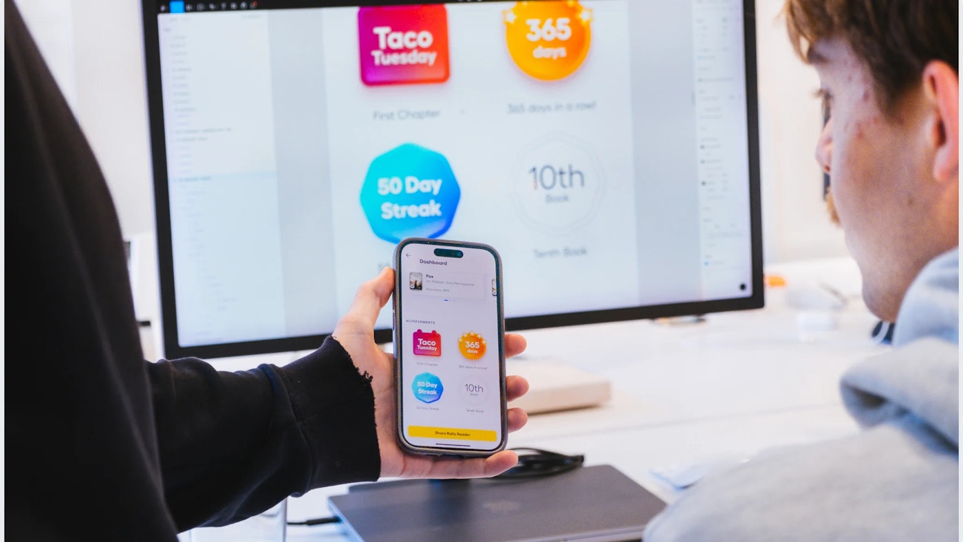 Hand holding an iPhone that shows an interface with bright colorful badges. In the background, slightly out of focus, is a monitor that shows the very same interface on a large scale.