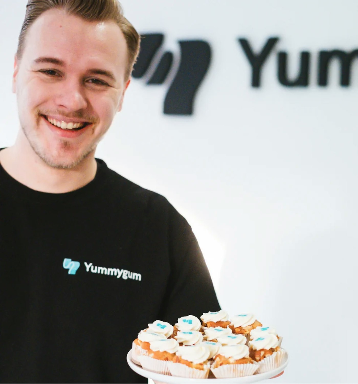 Person smiling at camera holding a plate with small cupcakes that each have a turquoise Yummygum logo mark on top. Person is wearing a near black sweater that shows the same logo mark accompanied by the word Yummygum shown in white. In the back is a white wall that shows a small part of a dark logo.