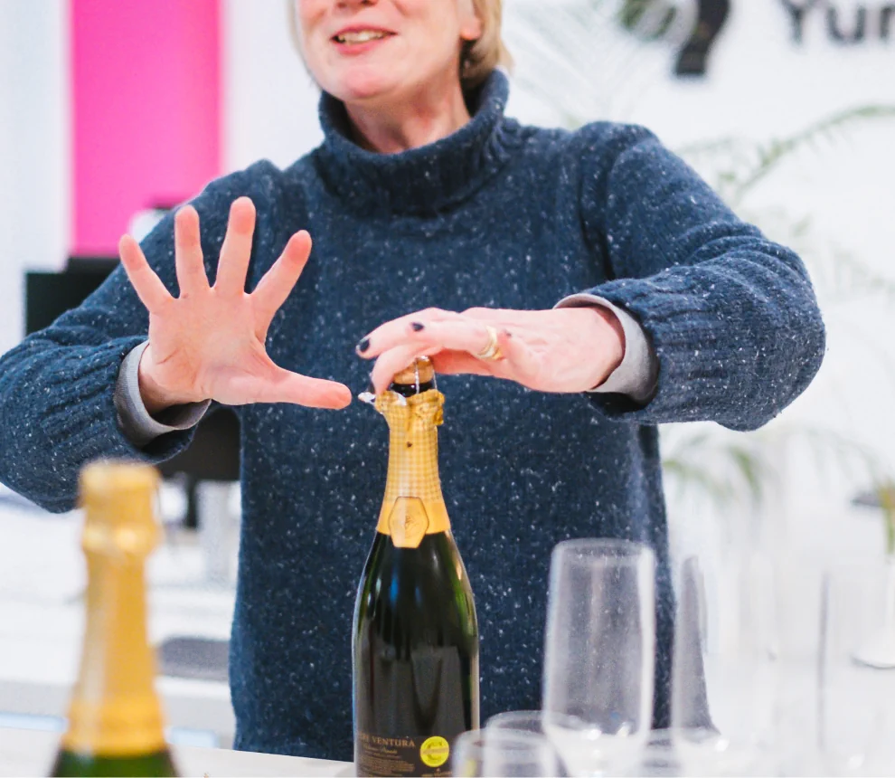 A person's with a knitted sweater opening a champagne bottle with two hands of which one make expressive gesture.  The face of the person is just out of crop.