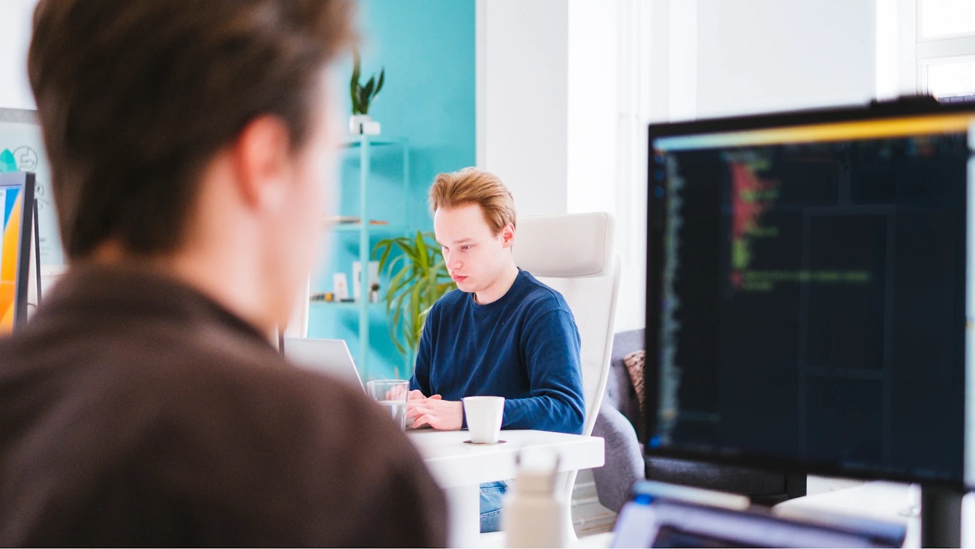 One person who's working behind their laptop in focus as seen in between a second out-of-focus person and their monitor.