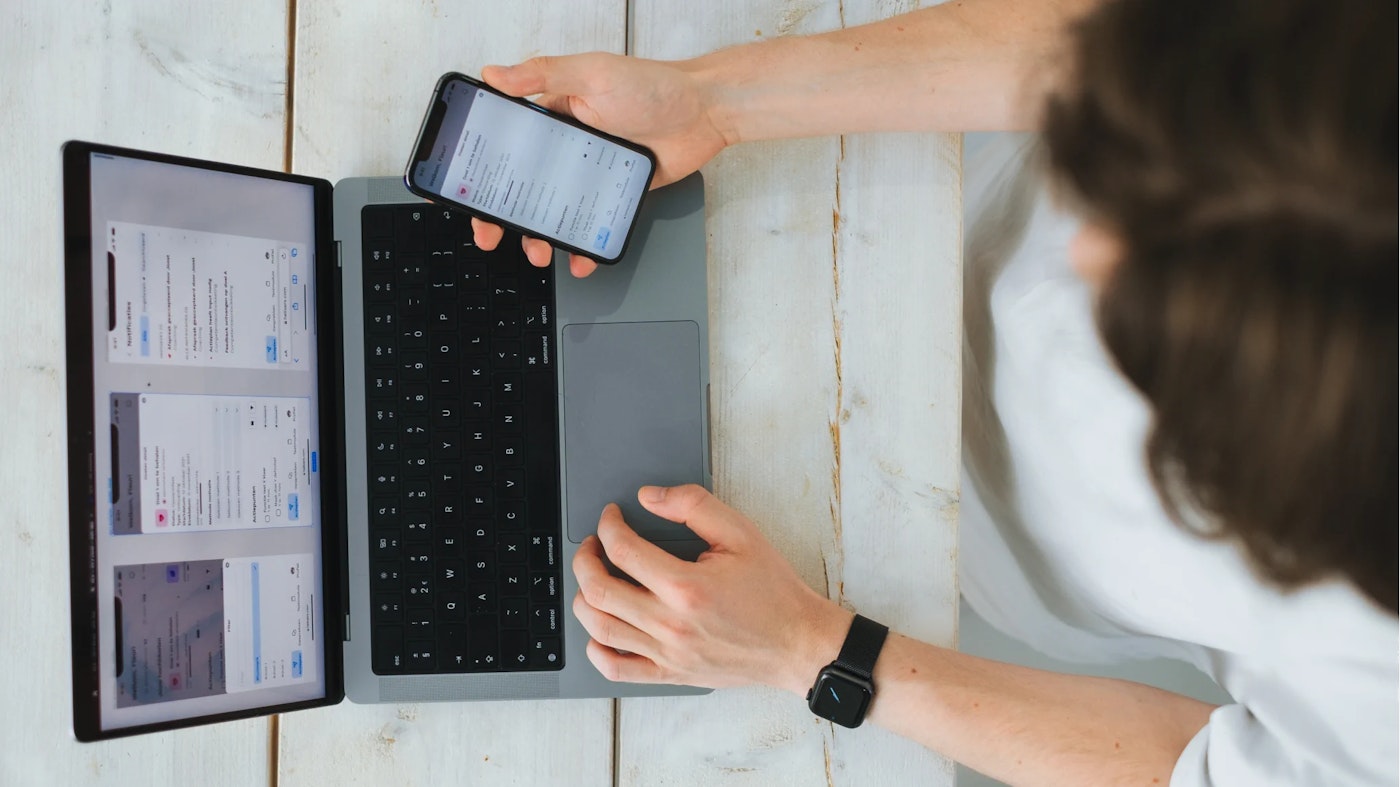 Person as seen from above, holding iPhone right next to their laptop screen. Both the laptop screen and the iPhone screen shot a mainly white and minimalist interface with dashes of color.