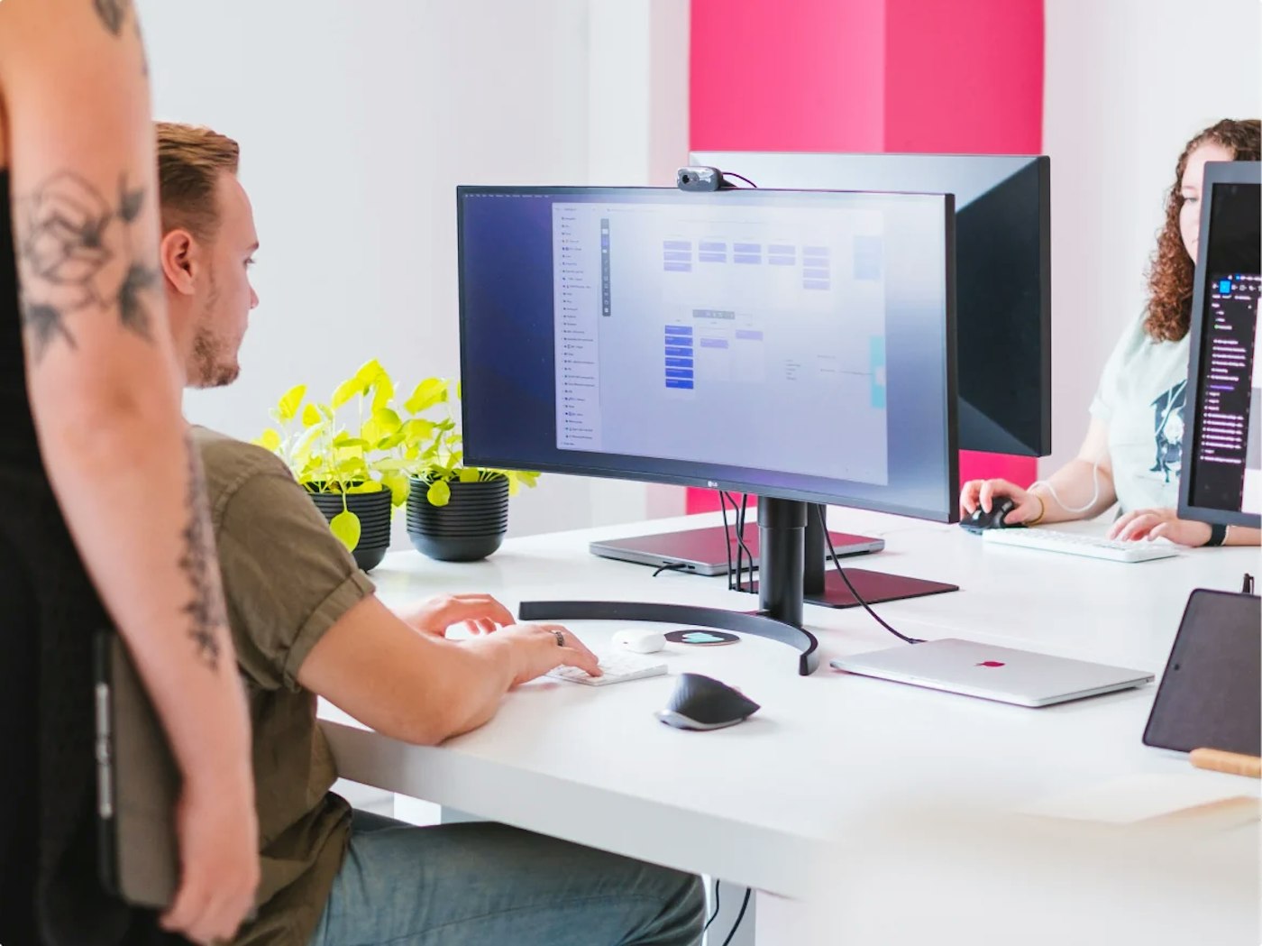 Person typing while looking at computer screen that is standing on a white desk. Someone is standing next to him. Only this person's arm shows. In the back is someone else working on their computer.