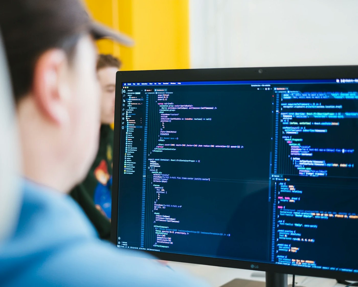 Out of focus person looking at a, in-focus, computer monitor that shows a dark blue code editor. In the rear is a bright yellow colored wall.