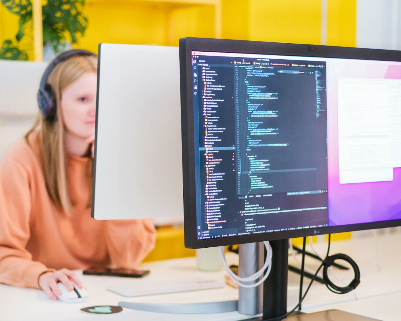 A desk with a computer monitor showing a code editor. In the rear is a person wearing an orange sweater with headphones on, as seen from the front, working behind another computer monitor.