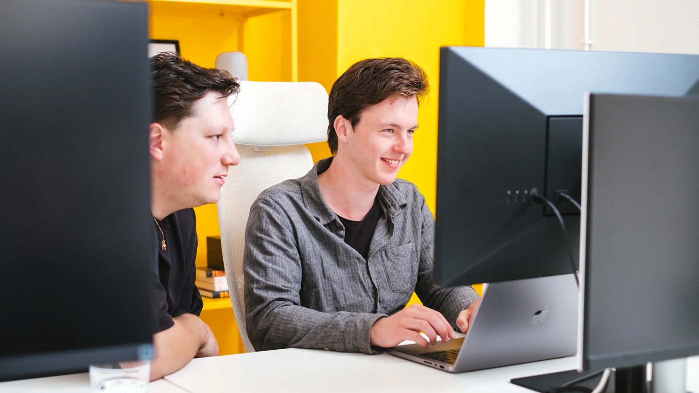 Two people sitting side by side behind a set of computer monitors. One of them is looking at the other, while the other person is smiling..